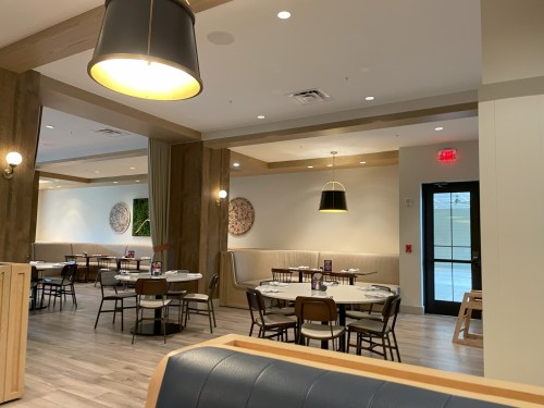 Modern restaurant interior with wooden accents, tables set for dining, and large windows letting in natural light.