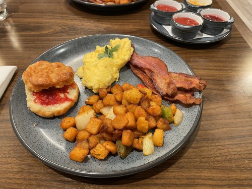 A plate with scrambled eggs, bacon, a biscuit with jam, and seasoned potato cubes, alongside small bowls of sauces.