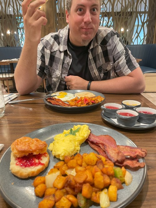 A man sits at a table with a hearty breakfast, including eggs, bacon, potatoes, and biscuits with jam.
