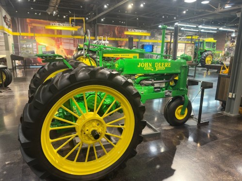 A vintage John Deere tractor displayed in a museum, featuring green and yellow colors, with a modern exhibit backdrop.