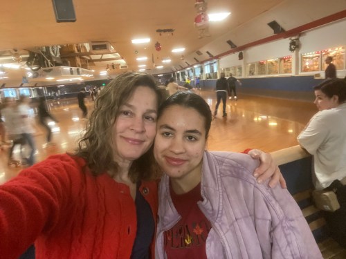 Two women smile for a selfie at a roller skating rink, with skaters in the background.