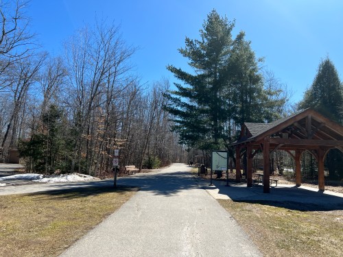 A clear path leads through a wooded area, with a shelter on the right and snow patches on the ground.