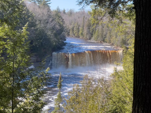 A scenic view of a waterfall surrounded by lush green trees and a flowing river under a clear blue sky.