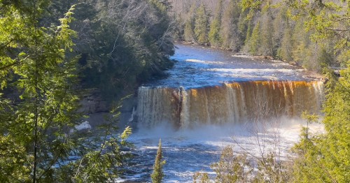 A scenic view of a waterfall cascading into a river, surrounded by lush green trees and a clear blue sky.