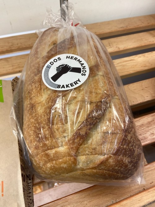 A loaf of bread in a clear bag with a "Dos Hermanos Bakery" label on a wooden shelf.