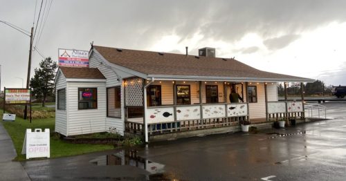 A small, white restaurant with a brown roof, open sign, and fish decorations, set against a cloudy sky.