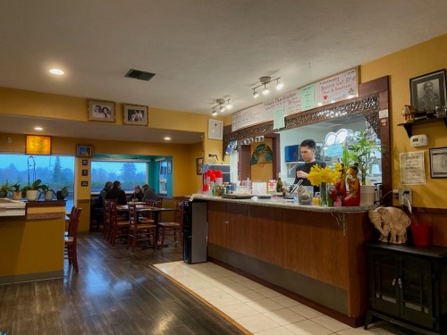 A cozy restaurant interior with a counter, plants, and a customer being served, featuring warm lighting and wooden decor.