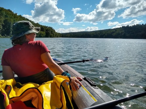 A person in a hat sits in a kayak on a calm lake, surrounded by trees and under a blue sky with fluffy clouds.