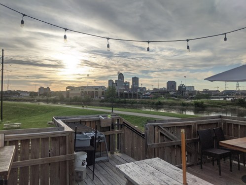 Sunset view of a city skyline from a wooden deck, with string lights and a river in the foreground.