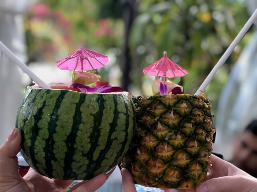 Two tropical drinks served in carved watermelon and pineapple, each adorned with pink umbrellas and flowers.