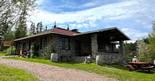 A stone building with a red roof, surrounded by trees and a gravel path, featuring a picnic table in the foreground.