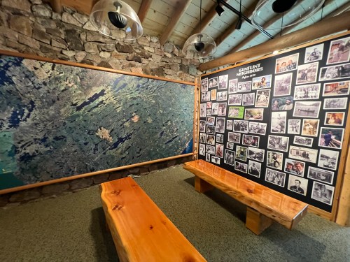 A room with a stone wall featuring a large map and a collage of black-and-white photos, with wooden benches in front.