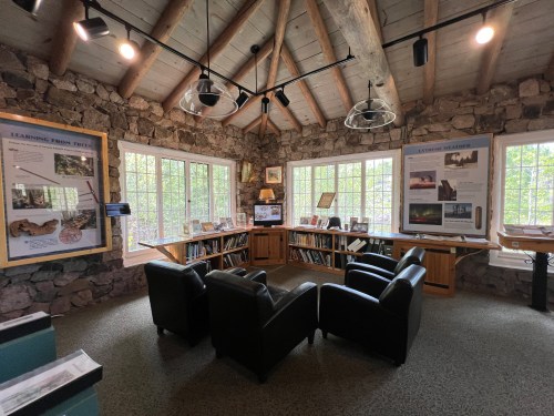 A cozy room with stone walls, large windows, and two black chairs, featuring informational displays and bookshelves.