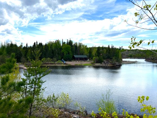 A serene lake surrounded by lush greenery and trees, with a cabin visible on the shore under a cloudy sky.