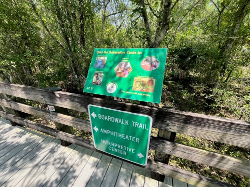 Signpost directing visitors to the boardwalk trail, amphitheater, and interpretive center in a lush, green environment.
