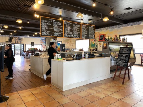 A bright café interior with a counter, menu boards, and a staff member serving customers. Tables are set for dining.