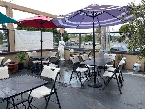 Outdoor seating area with tables and chairs under colorful umbrellas, featuring a statue and a view of the street.