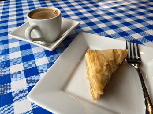 A slice of baklava on a white plate next to a small cup of coffee, set on a blue and white checkered tablecloth.