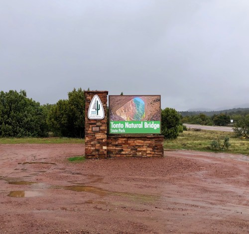 Sign for Tonto Natural Bridge State Park, surrounded by greenery, on a cloudy day.