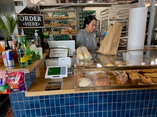 A woman at a bakery counter prepares an order, with pastries and drinks displayed behind her.