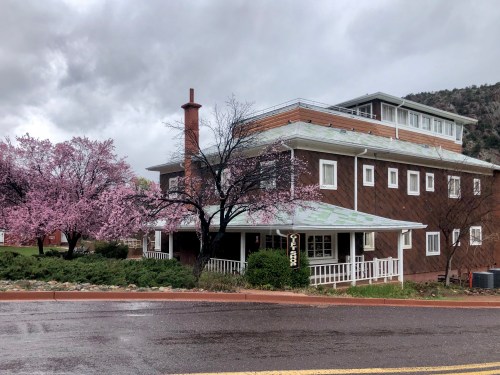 A brown building with a white porch, surrounded by blooming pink trees, under a cloudy sky.