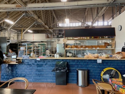 A cozy bakery interior with a blue tiled counter, shelves of baked goods, and a staff member preparing orders.