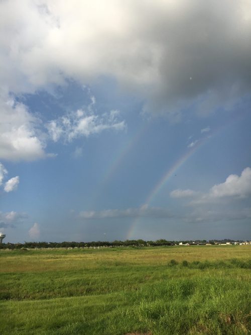A vibrant double rainbow arcs over a green field under a partly cloudy sky.