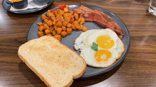 A plate with two fried eggs, crispy bacon, seasoned potatoes, and a slice of toasted bread on a wooden table.
