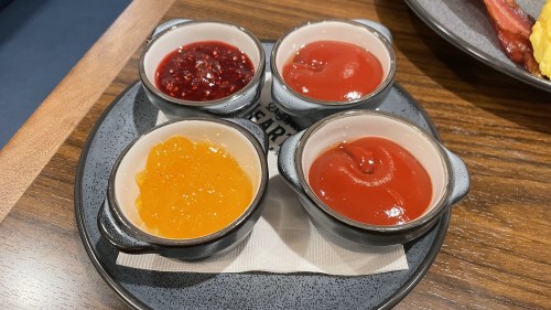 Four small bowls of different sauces: orange marmalade, red jam, ketchup, and another red sauce, on a plate.