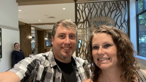 A smiling couple poses for a selfie in a modern lobby with wooden decor. A staff member is seen in the background.