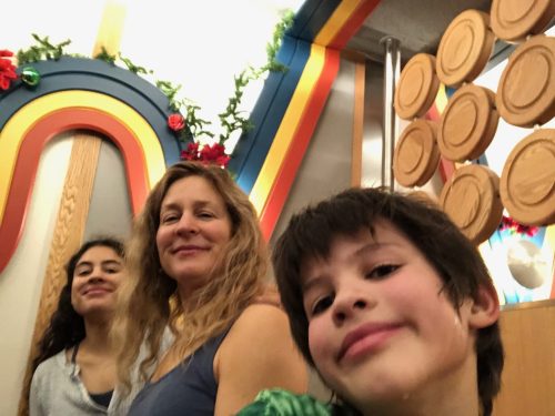 A woman and two children smile for a selfie in a colorful, festive setting with decorative elements in the background.