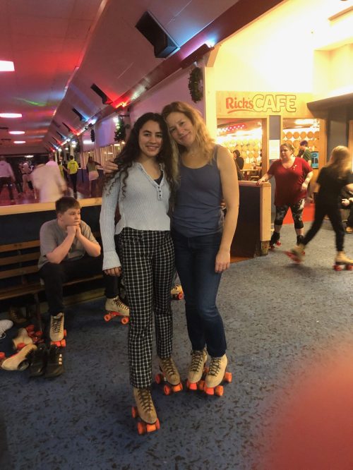 Two women pose together in a roller skating rink, wearing skates, with colorful lights and people skating in the background.