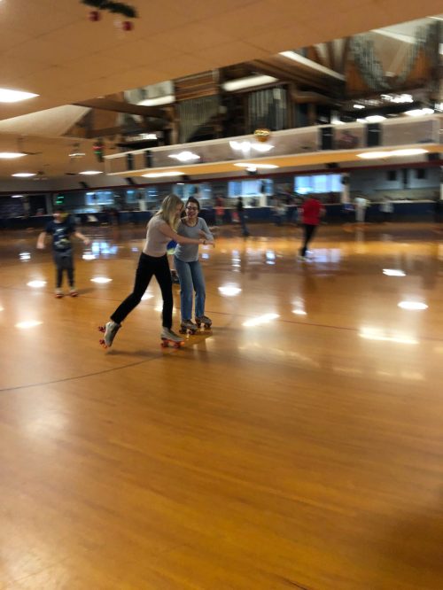 Two people skate together in a roller rink, surrounded by others enjoying the activity on a polished wooden floor.