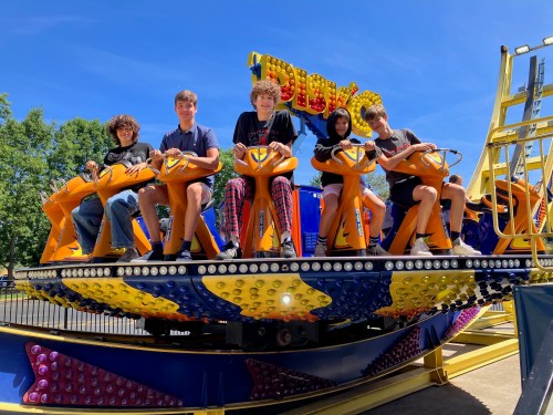 A group of five friends sits on a colorful amusement park ride, smiling under a clear blue sky.