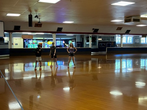 Three children stand on a shiny roller skating rink, with arcade games visible in the background.