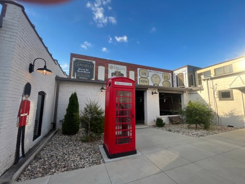 A red telephone booth stands outside a white brick building with vintage signs and a clear blue sky.