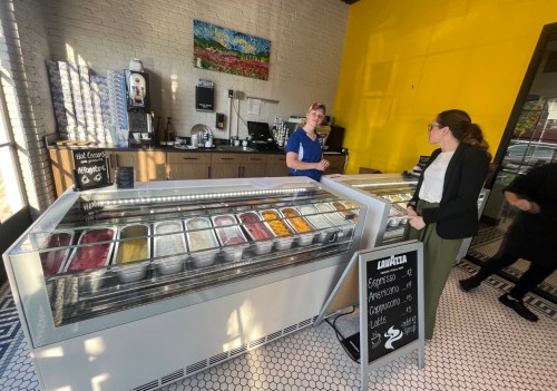 A woman in a blue shirt serves gelato at a counter, while a customer in a black blazer looks at the selection.