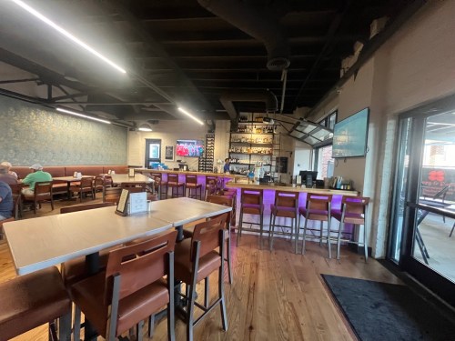 Interior of a restaurant featuring a bar, seating area, and a TV, with warm lighting and wooden floors.