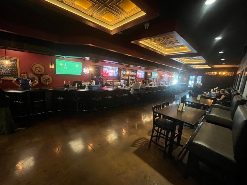 A spacious bar interior with a long counter, tables, and screens showing sports, illuminated by warm lighting.
