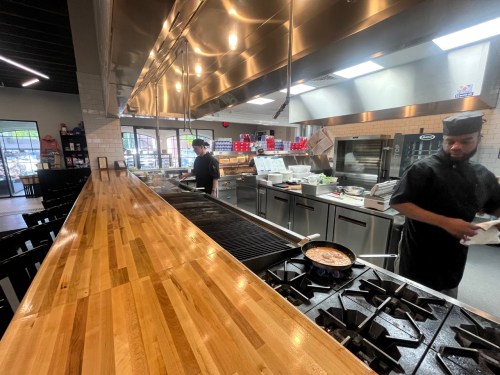 A modern kitchen with chefs cooking, featuring a grill, stovetop, and a long wooden counter. Bright and spacious atmosphere.
