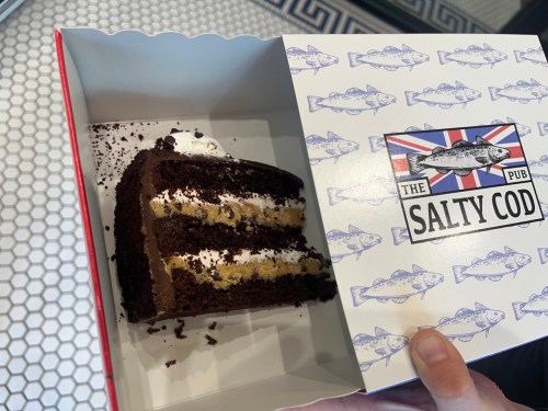 A slice of chocolate cake with cream and a cookie layer, in a box labeled "The Salty Cod."