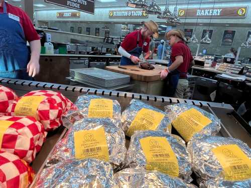 Two workers in red shirts and cowboy hats prepare food at a counter, with wrapped items displayed in the foreground.