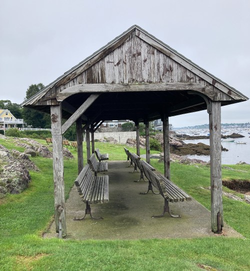 A weathered wooden shelter with benches overlooks a rocky shoreline and calm water on a cloudy day.
