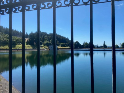 A serene lake view framed by a black metal fence, surrounded by trees and a clear blue sky.