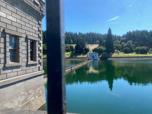 A serene view of a reservoir with a stone building and lush greenery reflected in the calm water under a clear blue sky.