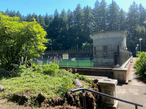 A stone structure surrounded by greenery and trees, with a fenced area and clear blue sky above.