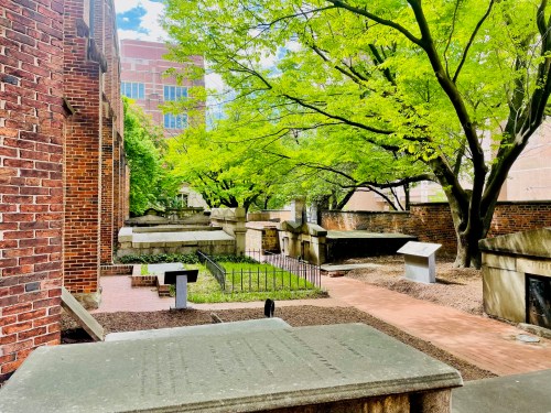 A peaceful cemetery with stone graves surrounded by lush green trees and brick buildings in the background.