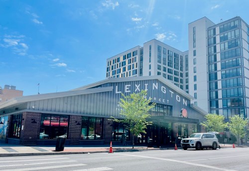 Lexington Market building with modern architecture, blue sky, and trees in front. Cars parked on the street.