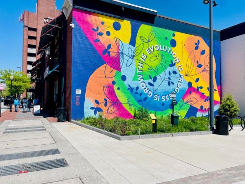 Colorful mural on a building featuring vibrant floral designs and the text "Growth is Evolution's Space." Sidewalk and people nearby.