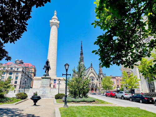 A sunny city square featuring a tall monument, a statue, and a historic church surrounded by greenery and parked cars.
